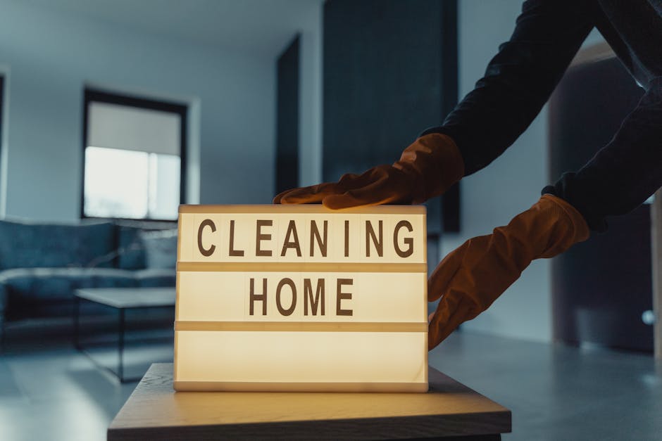 Close-up of a person wearing black clothing and brown rubber gloves adjusting a small lightbox sign that reads 'CLEANING HOME' on a wooden table in a modern living room with a dark sofa, a coffee tabl